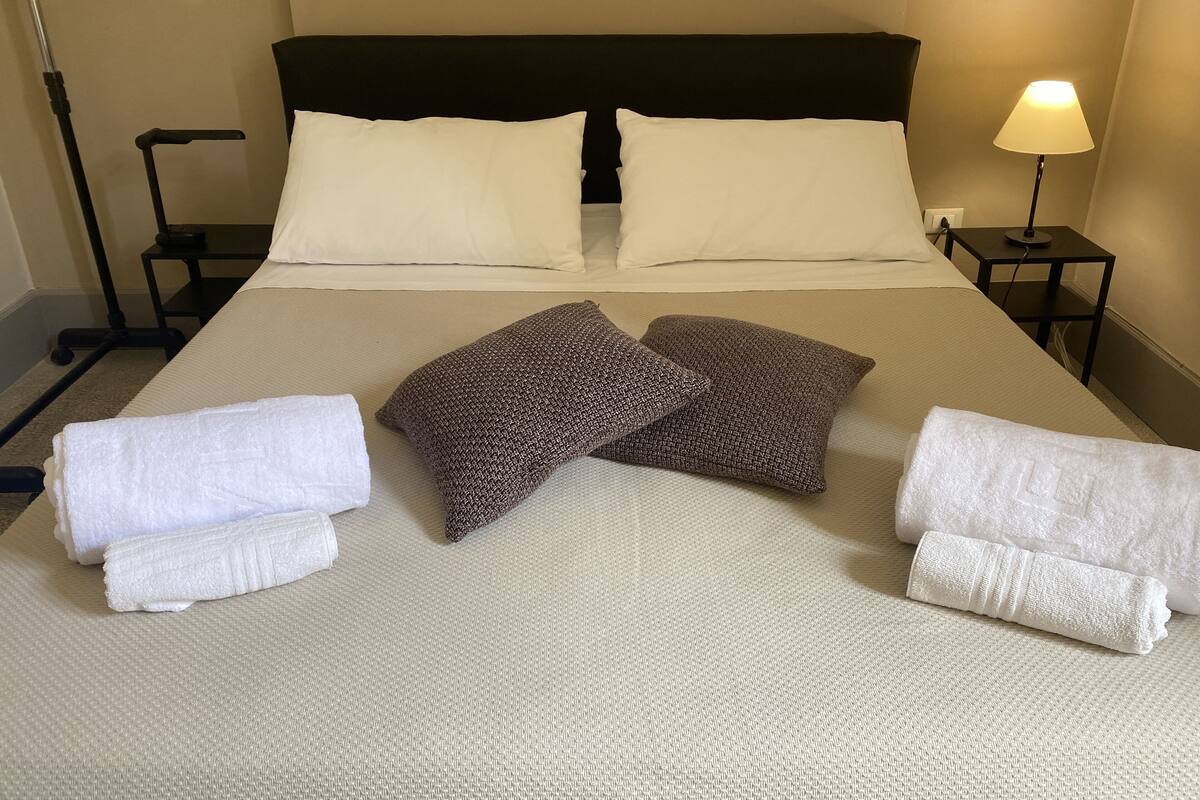 Een net opgemaakt bed met witte en bruine kussens, een opgevouwen witte handdoek en een lamp op een nachtkastje.