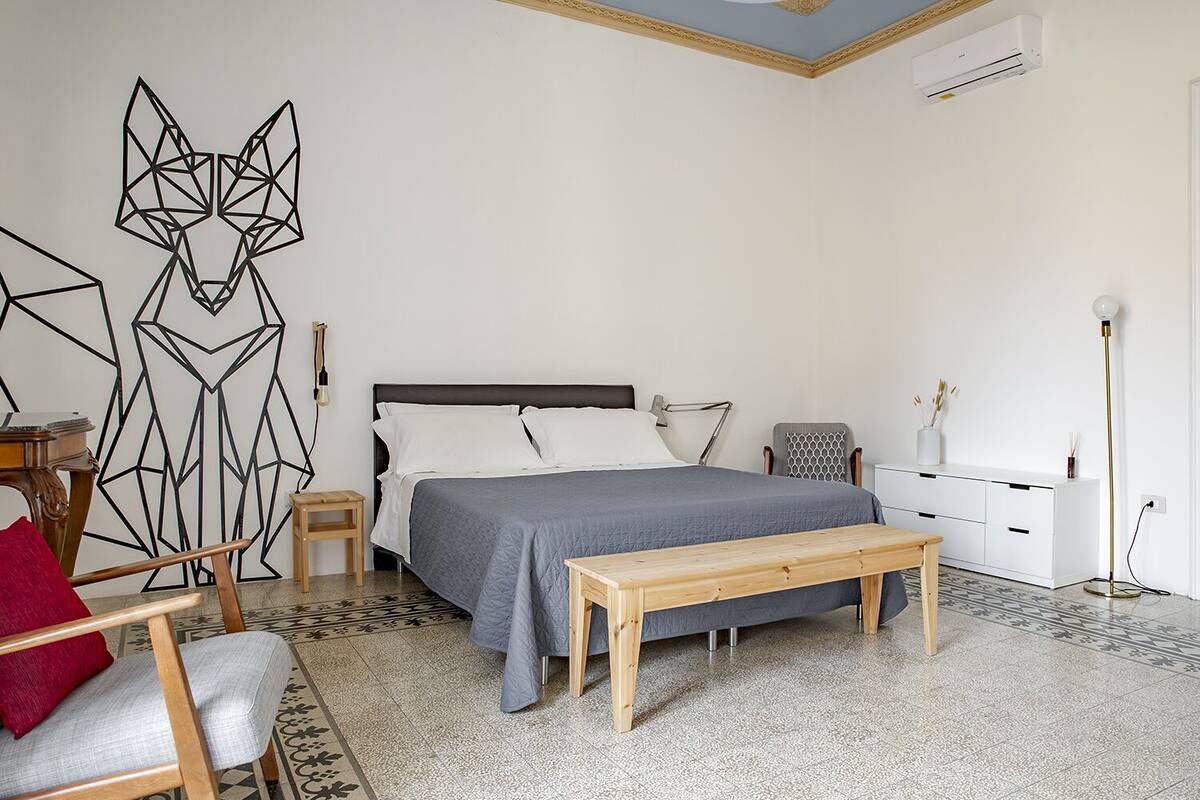Een slaapkamer met een bed, nachtkastjes, een bankje, een stoel, een dressoir en een wanddecoratie met geometrische patronen.
