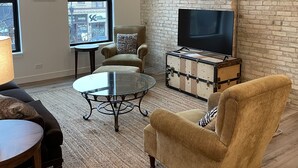 A living room with a brick wall, a ceiling fan, a television on a wooden chest, and a sofa. - 15 minutes to EAA. Newly renovated, Luxury 1 Bedroom in historic downtown Neenah (Neenah)