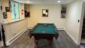 Una mesa de billar en una habitación con suelo de madera, paredes blancas y una ventana con persianas.