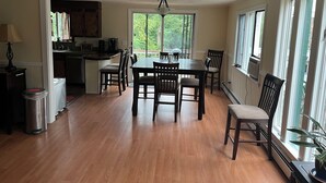 Un comedor amplio con una mesa y sillas de madera, una zona de cocina con armarios y encimera, y ventanas grandes que ofrecen luz natural.