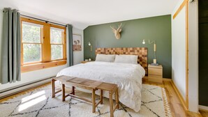 Una camera da letto con una testiera in legno, un'ampia finestra con tende, un comodino e una panca.