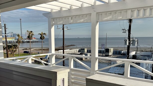 A covered deck with a view of the beach and ocean, featuring a wooden floor and white railings.