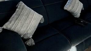 A dark blue sofa with a knitted throw blanket and a decorative pillow, facing a large window with a view of a beach, palm trees, and amusement park rides.