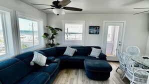 A modern living room with a dark blue sectional sofa, a glass-top dining table with white wicker chairs, a ceiling fan, and large windows offering a view of the beach.