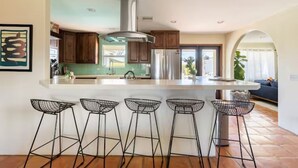 A modern kitchen with a large island, bar stools, wooden cabinets, and a view to the living area. - Casa Bermuda a Spanish Boho Retreat, Expansive Pool/Spa Pet Friendly (Bermuda Dunes)
