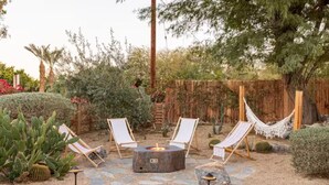 An outdoor seating area with a stone fire pit, surrounded by cacti and desert plants, with a wooden fence and hammock in the background. - Casa Bermuda a Spanish Boho Retreat, Expansive Pool/Spa Pet Friendly (Bermuda Dunes)