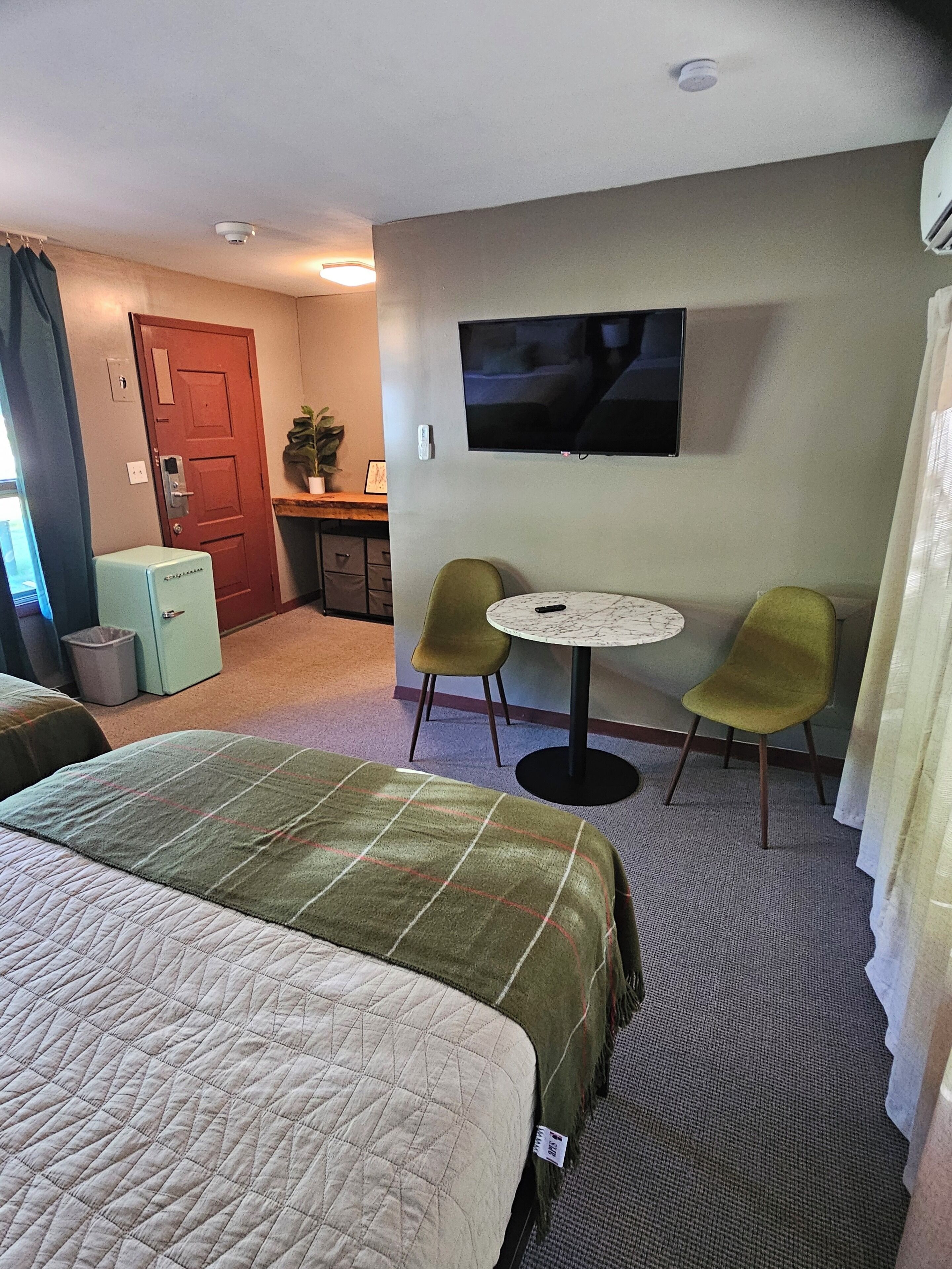 Habitación de hotel con dos camas, una mesita redonda pequeña con dos sillas, un televisor montado en la pared y un escritorio con una silla y una planta.