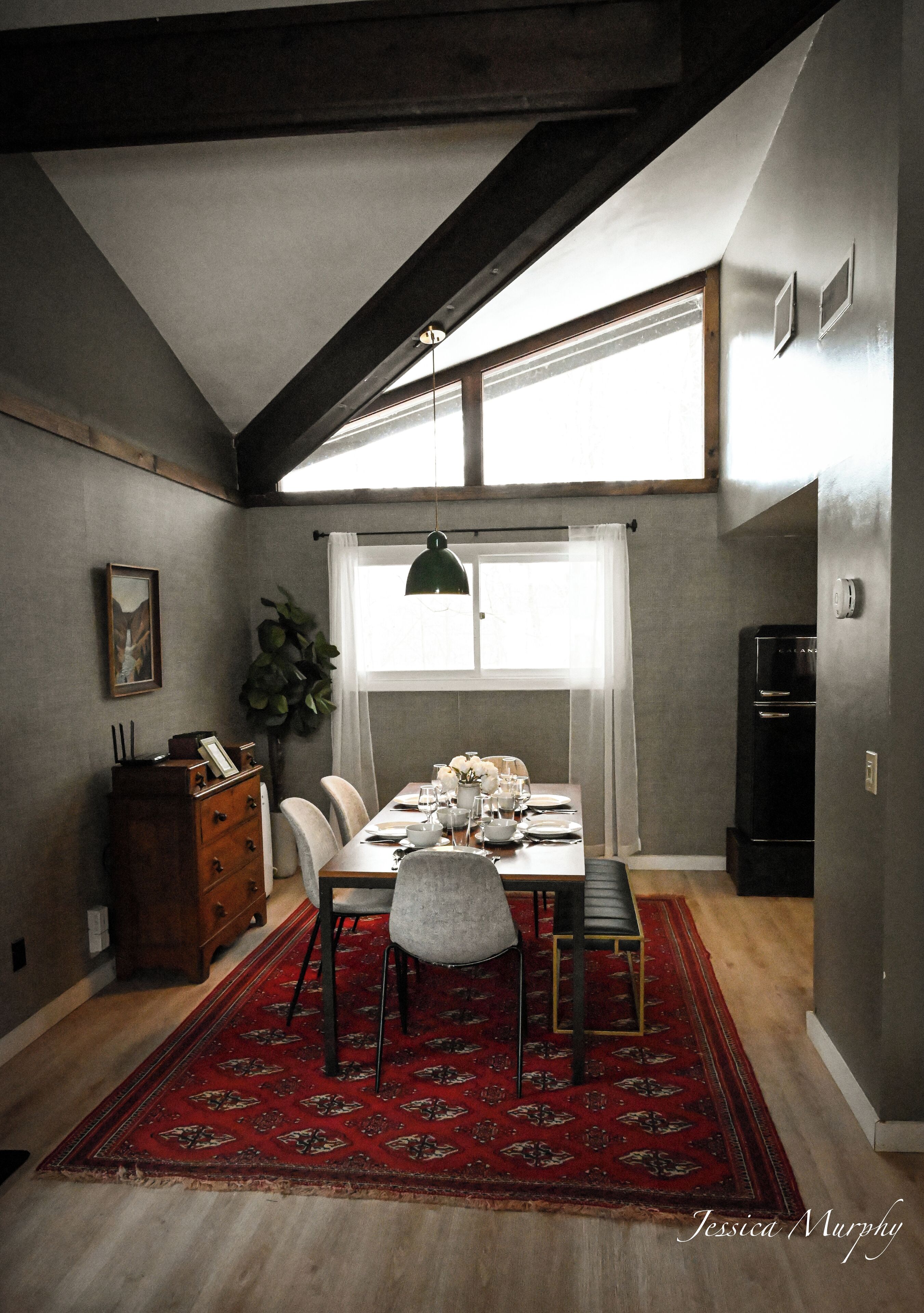 A dining area with a wooden table, chairs, a red patterned rug, a chandelier, and a large window with curtains.