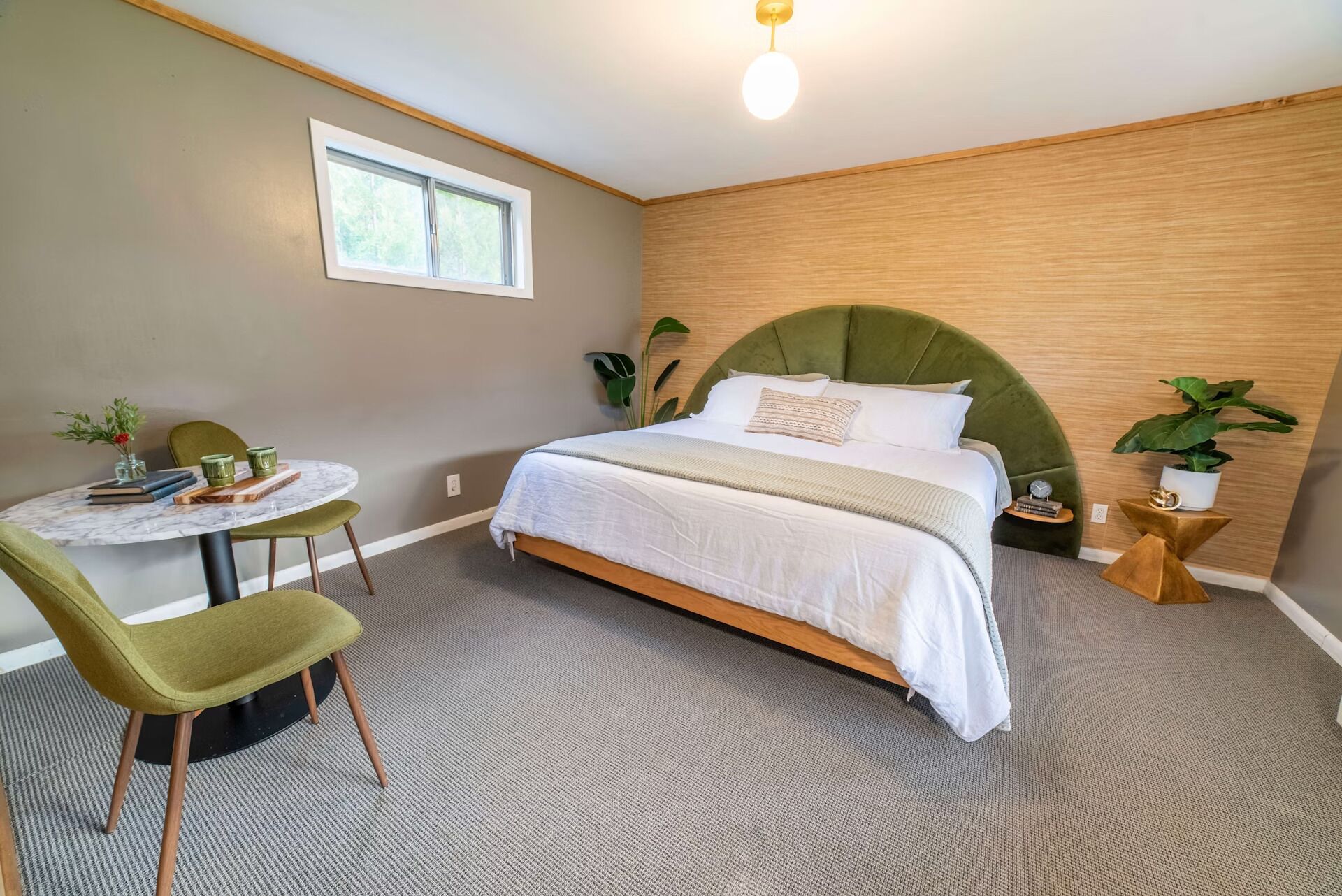 A bedroom with a bed, a round table with chairs, a window, and a plant.