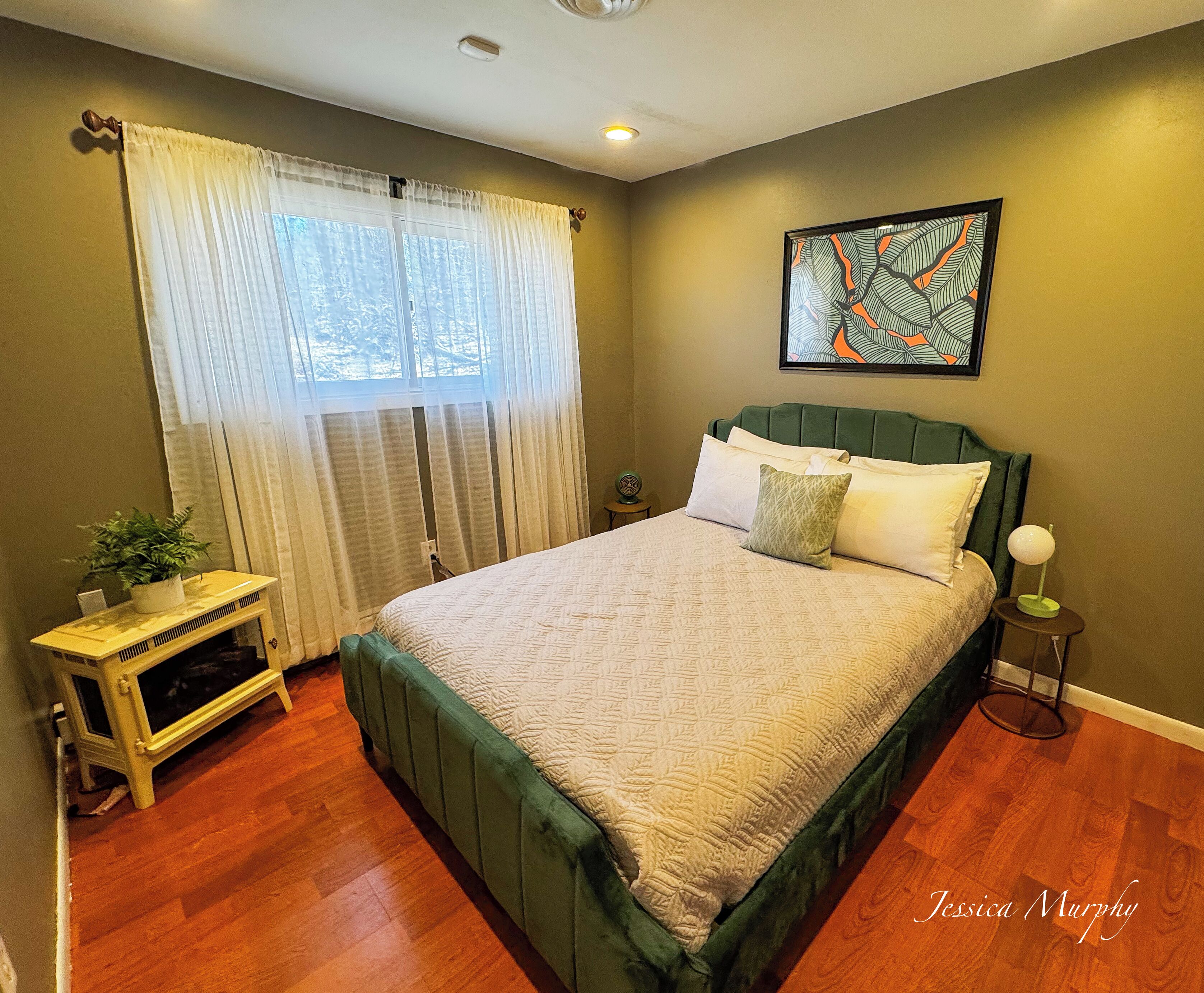 A bedroom with a green upholstered bed, a bedside table with a lamp, a small potted plant, and a window with sheer curtains.