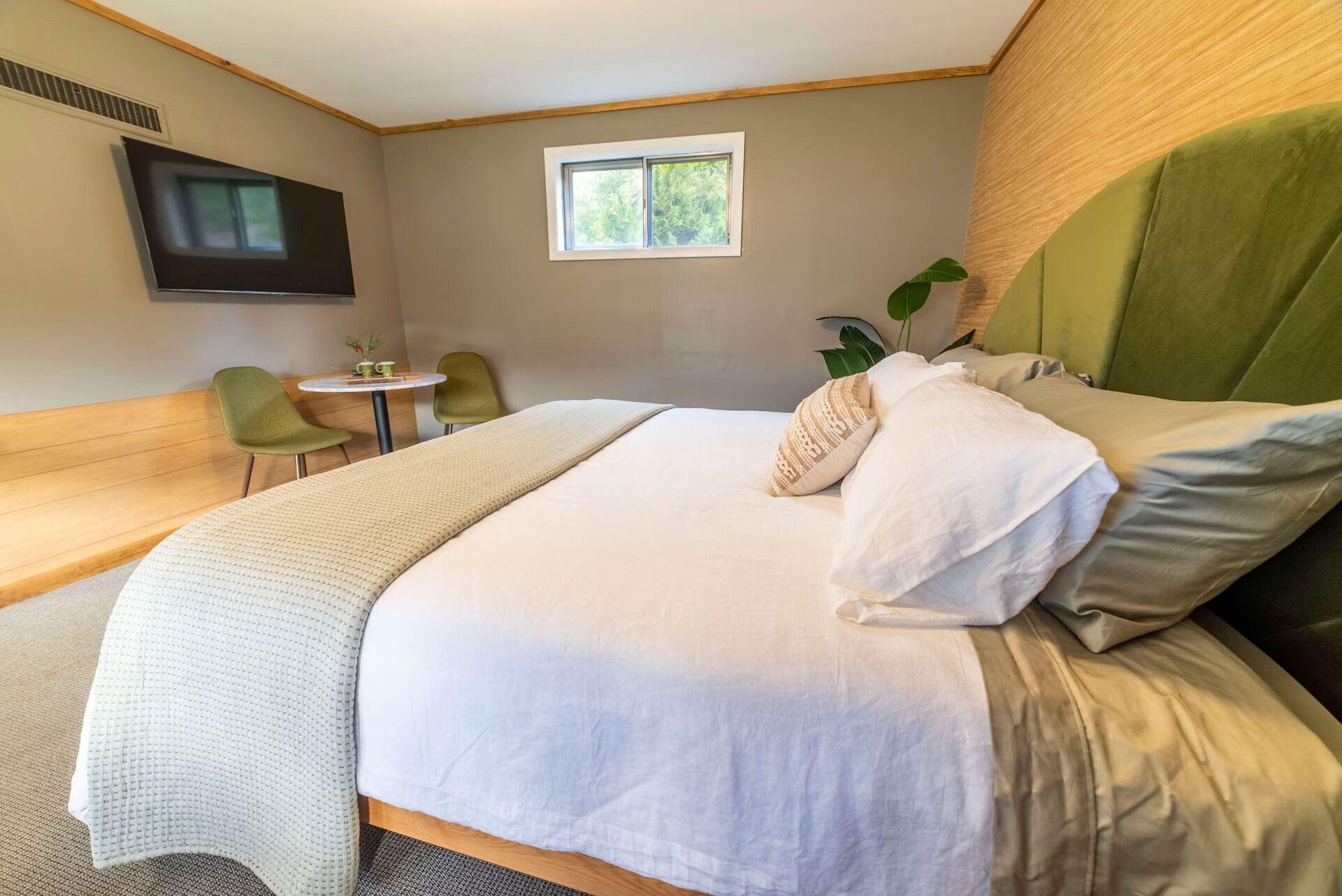Een hotelkamer met een groot bed, een aan de muur bevestigde televisie, een klein tafeltje met stoelen en een raam met uitzicht op groen.