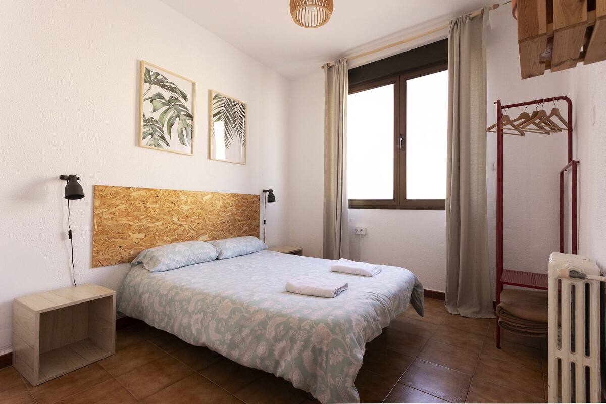 A bedroom with a wooden headboard, a bed with a patterned bedspread, two wall-mounted artworks, a window with curtains, and a radiator.