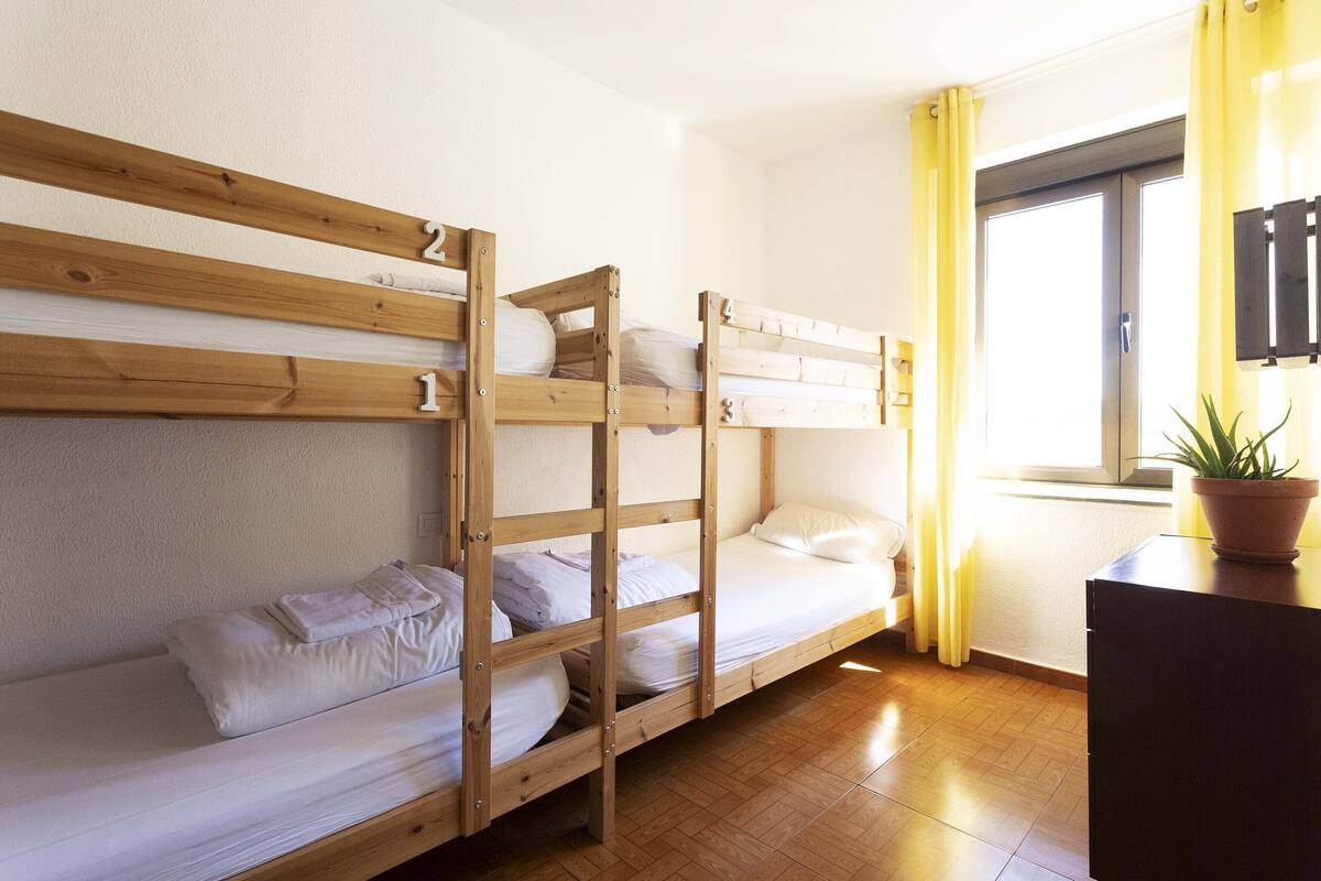 A bunk bed room with wooden frames, white bedding, and a potted plant on a desk.
