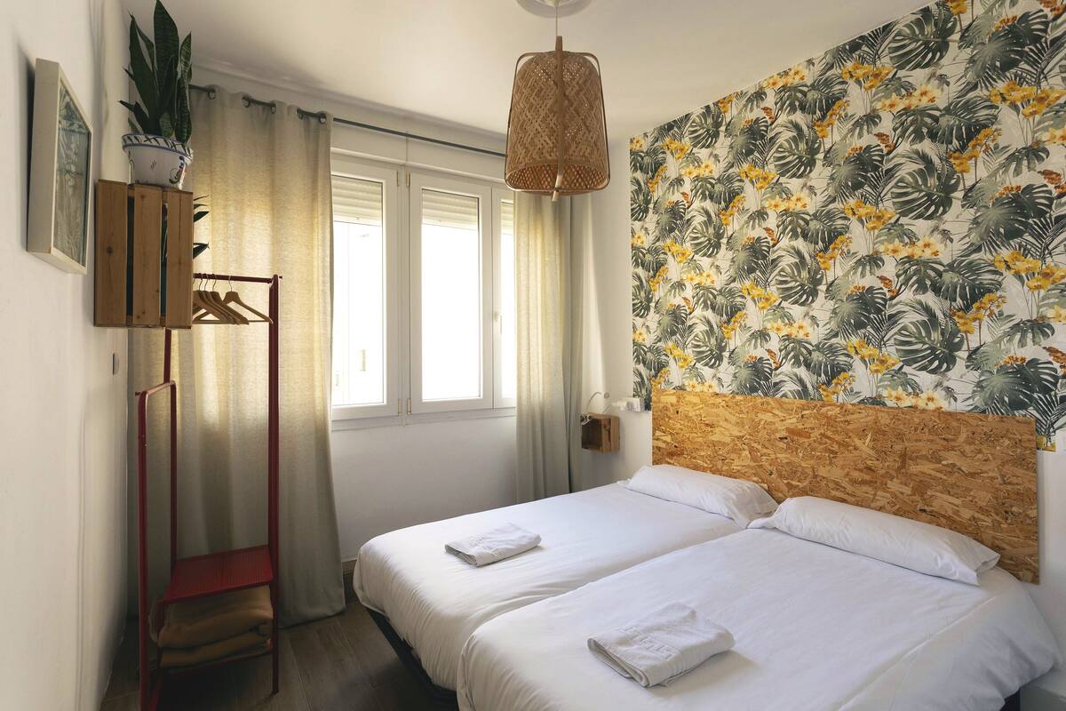 A hotel room with two beds, a wooden headboard, a tropical wallpaper, a hanging light fixture, and a plant on the wall.