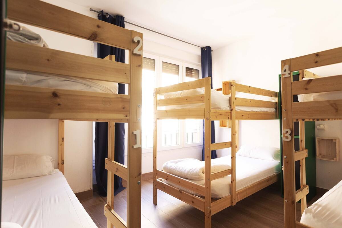 A dormitory-style room with bunk beds numbered 1 to 4, wooden walls, and a window with curtains.