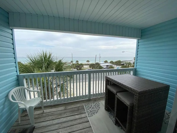 A covered balcony with a view of the beach, a white plastic chair, and a wicker table.