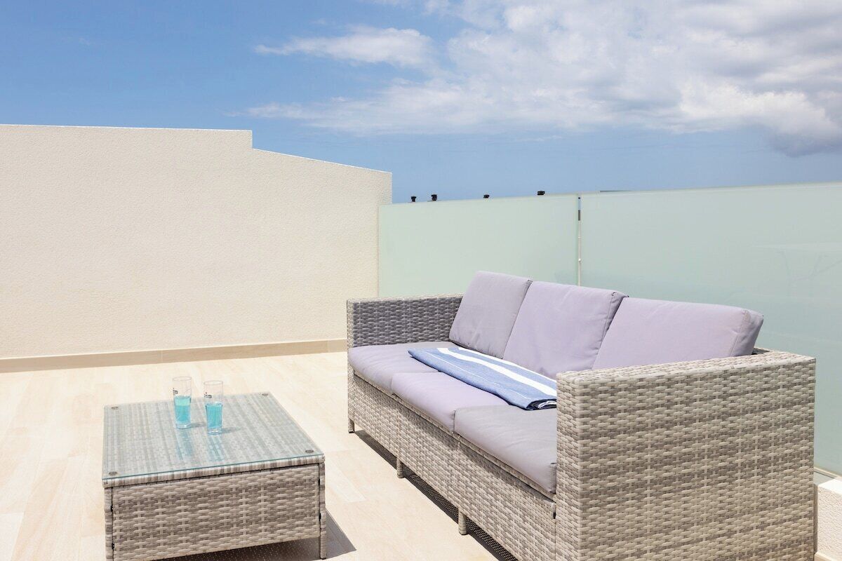 A balcony with a wicker sofa, a glass table with two glasses, and a light-colored wall.