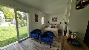 A bedroom with a bed, two blue armchairs, a wooden table, and a view of a garden through a sliding door.