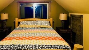 A bedroom with a sloped ceiling, a bed with patterned bedding, two bedside lamps, and a window with curtains.