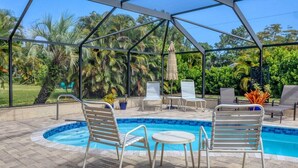 A covered outdoor swimming pool with lounge chairs and a table, surrounded by lush greenery.