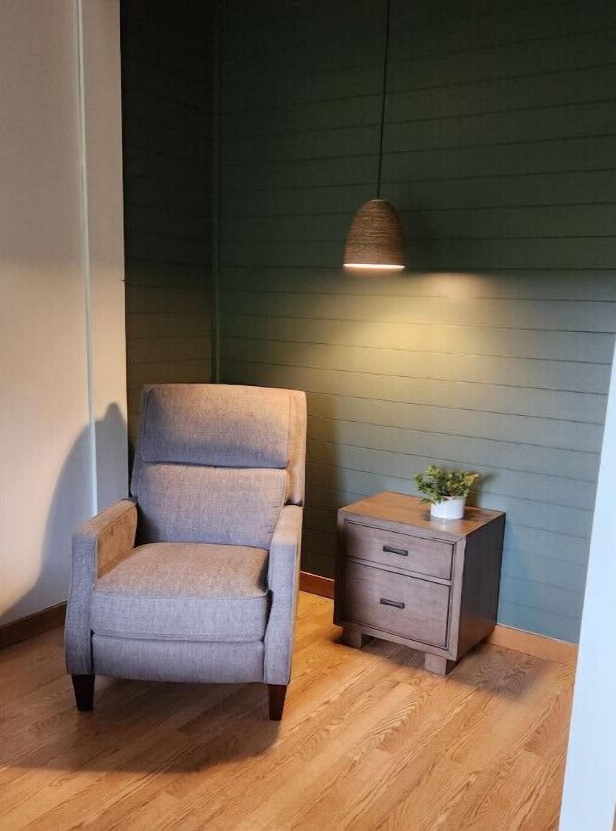 A room with a grey upholstered armchair, a wooden nightstand with drawers, and a hanging lamp.