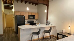 A compact kitchen with a refrigerator, microwave, and stove, a small bar area with two stools, and a bed in the adjacent room.