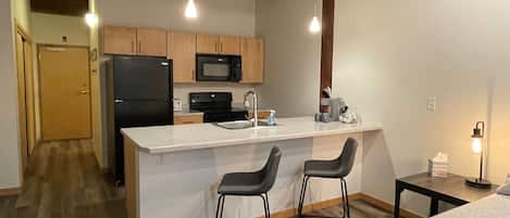 A compact kitchen with a refrigerator, microwave, and stove, a small bar area with two stools, and a bed in the adjacent room.
