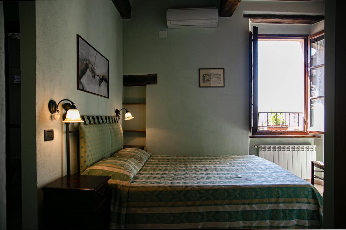 Une chambre à coucher comprenant un lit, deux lampes fixées au mur, un radiateur et une fenêtre donnant sur un paysage verdoyant.