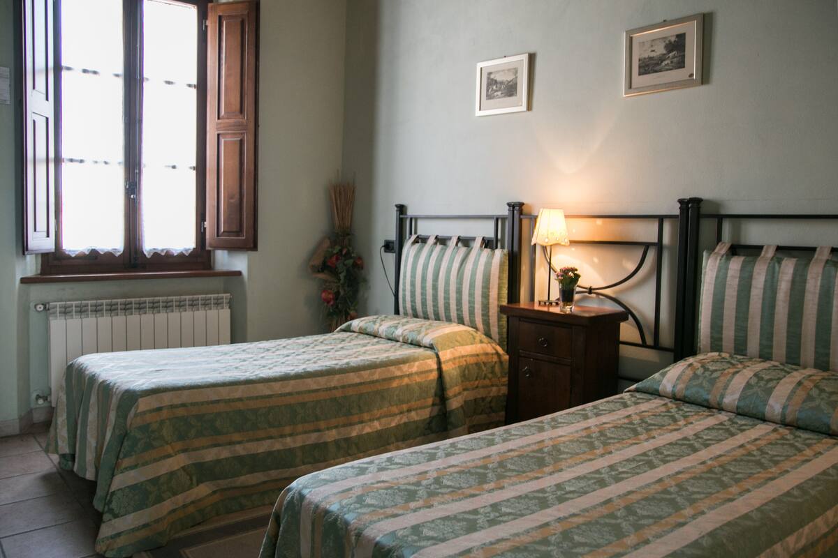 Une chambre avec deux lits simples, une table de chevet avec une lampe, un radiateur et une fenêtre dont les volets sont ouverts.