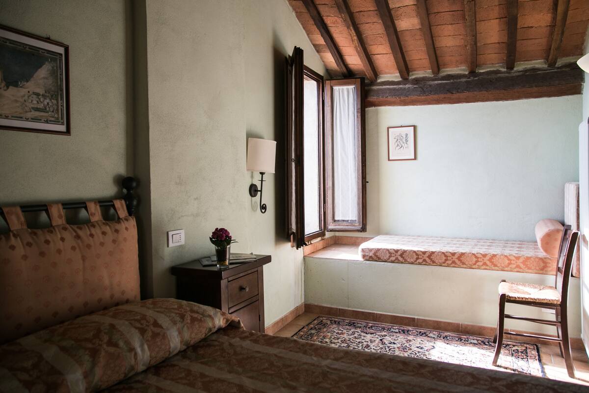Une chambre à coucher comprenant un lit, une chaise, une table de chevet, une fenêtre avec des rideaux et une lampe fixée au mur.