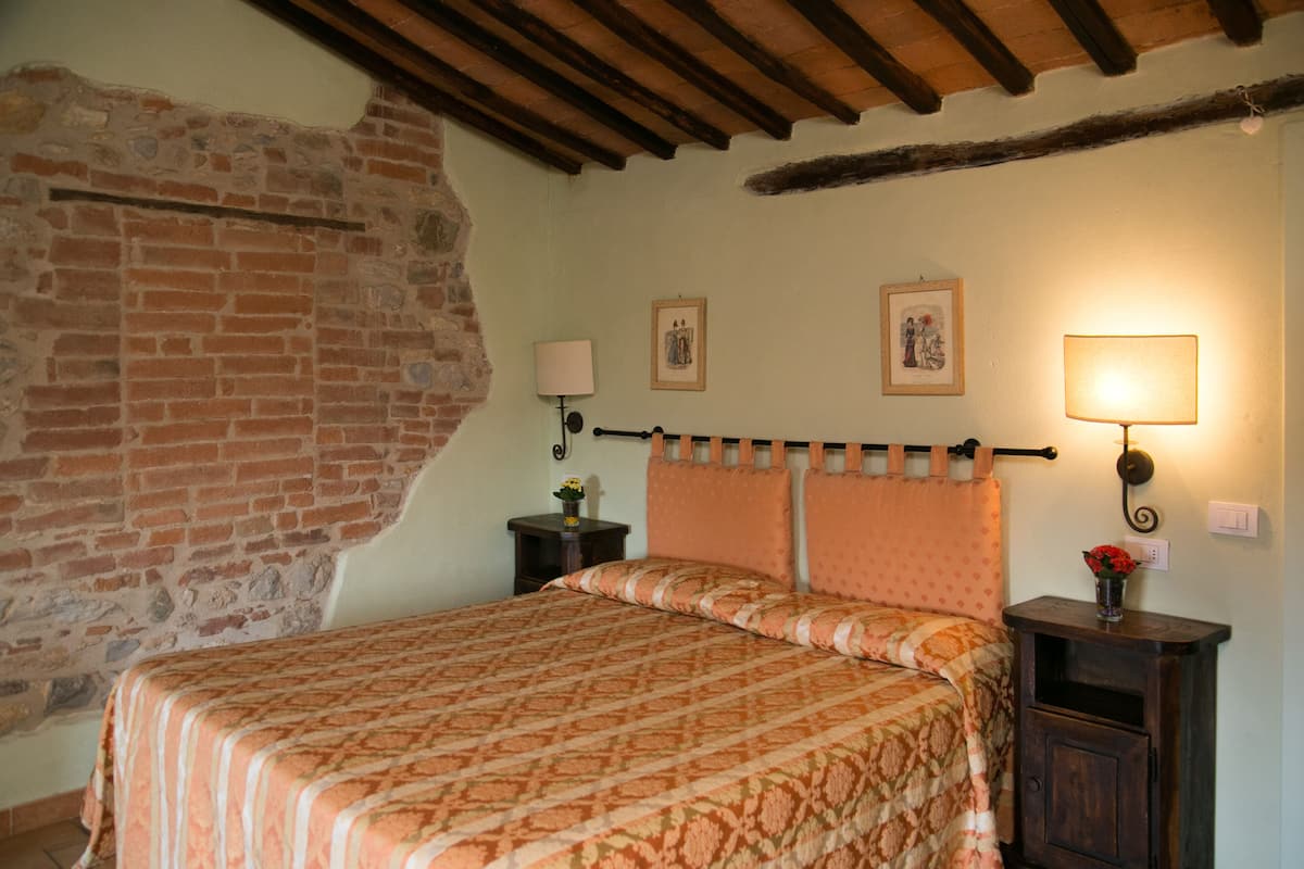 Une chambre avec un mur en briques, un lit recouvert d’une couvre-lit à motifs, deux lampes fixées au mur et une petite table sur laquelle est posé un vase de fleurs.