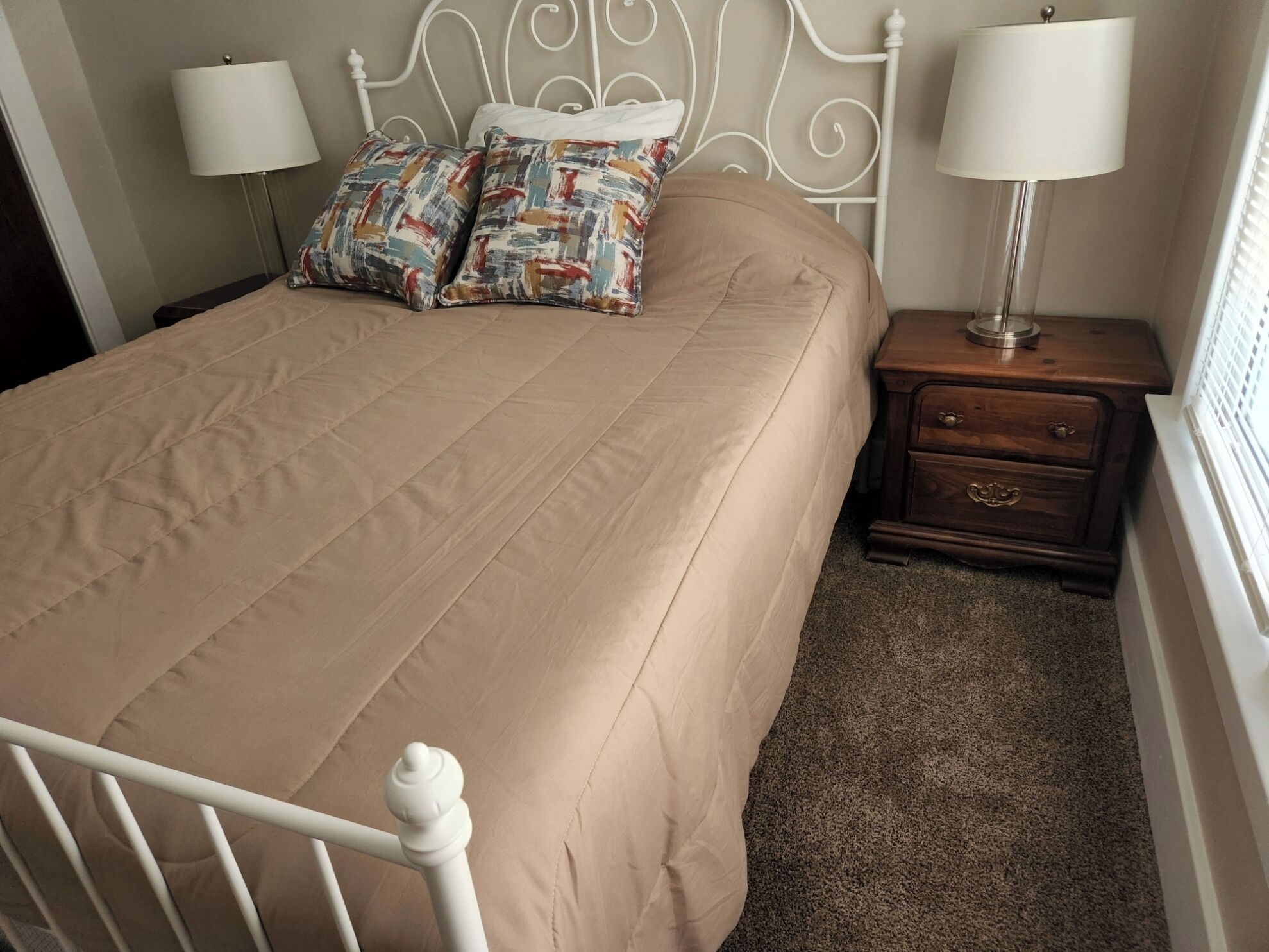 A neatly made bed with a beige bedspread and two colorful pillows, a white metal headboard, a wooden nightstand with a lamp, and a small drawer.
