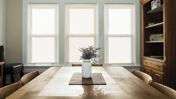 Un comedor con una gran mesa de madera, tres ventanas con persianas, un jarrón blanco con flores y un aparador de madera con estantes.