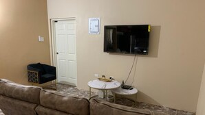 Een woonkamer met een bruine leren bank, een witte deur, een wandgemonteerde flatscreen televisie en twee kleine ronde tafels met decoratieve objecten.
