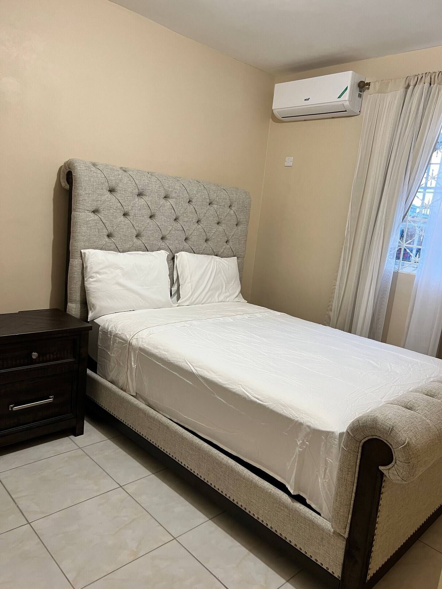 Une chambre à coucher avec une tête de lit capitonnée, un couvre-lit blanc, une table de chevet en bois foncé et un appareil de climatisation fixé au mur.