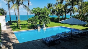 A swimming pool with lounge chairs and a striped umbrella, surrounded by palm trees and a view of the ocean. - Villa del Sol | 6BR | Exclusive Family Retreat (Cancún)