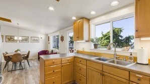 A modern kitchen with wooden cabinets, a large window, and a dining area with a table and chairs. - Experience Sequim Charm & Amazing Lighthouse Views, by the Olympic National Park (Sequim)
