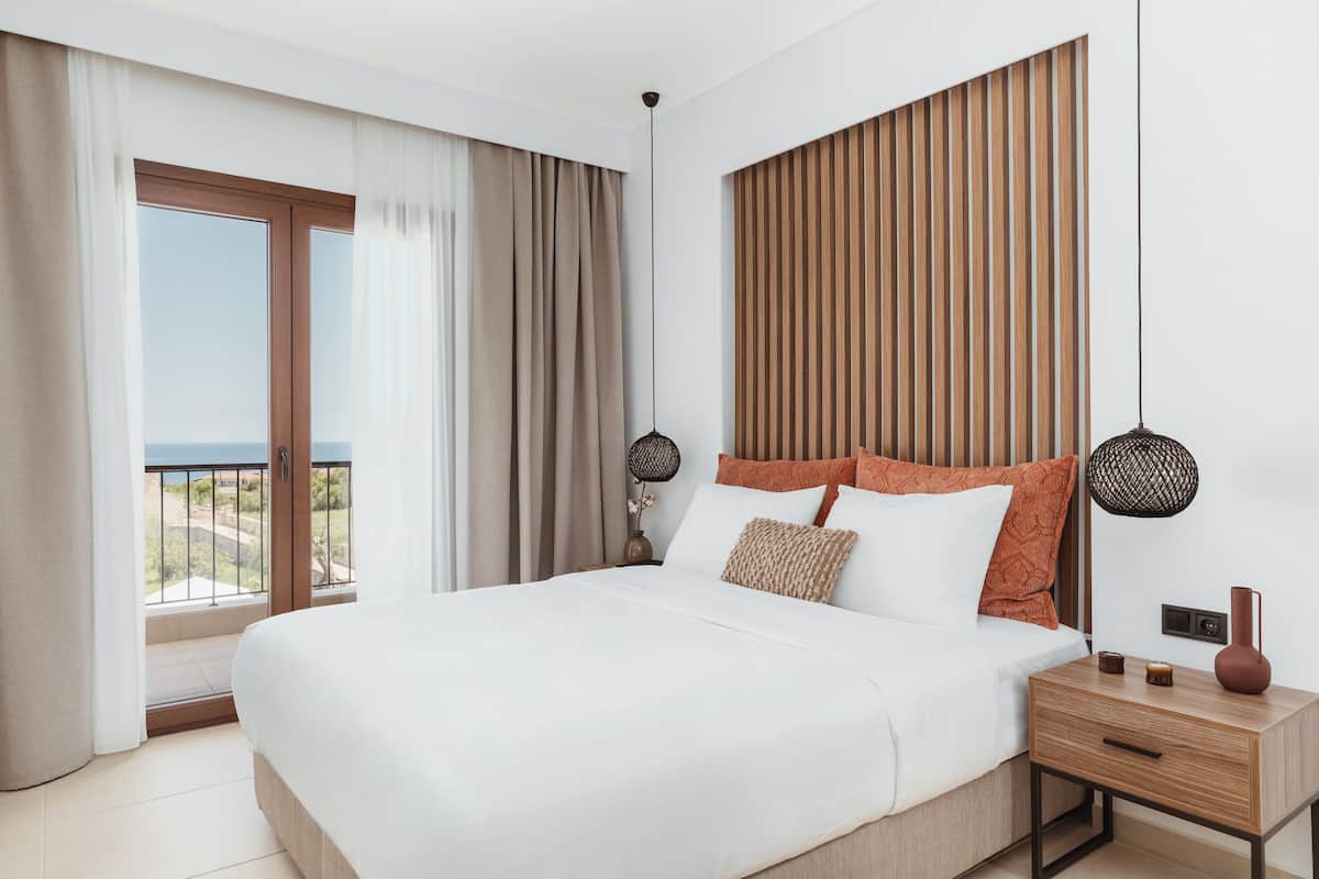 Habitación de hotel moderna con una cama grande, cabecera de madera, dos lámparas de noche, mesitas de noche y vistas al exterior.