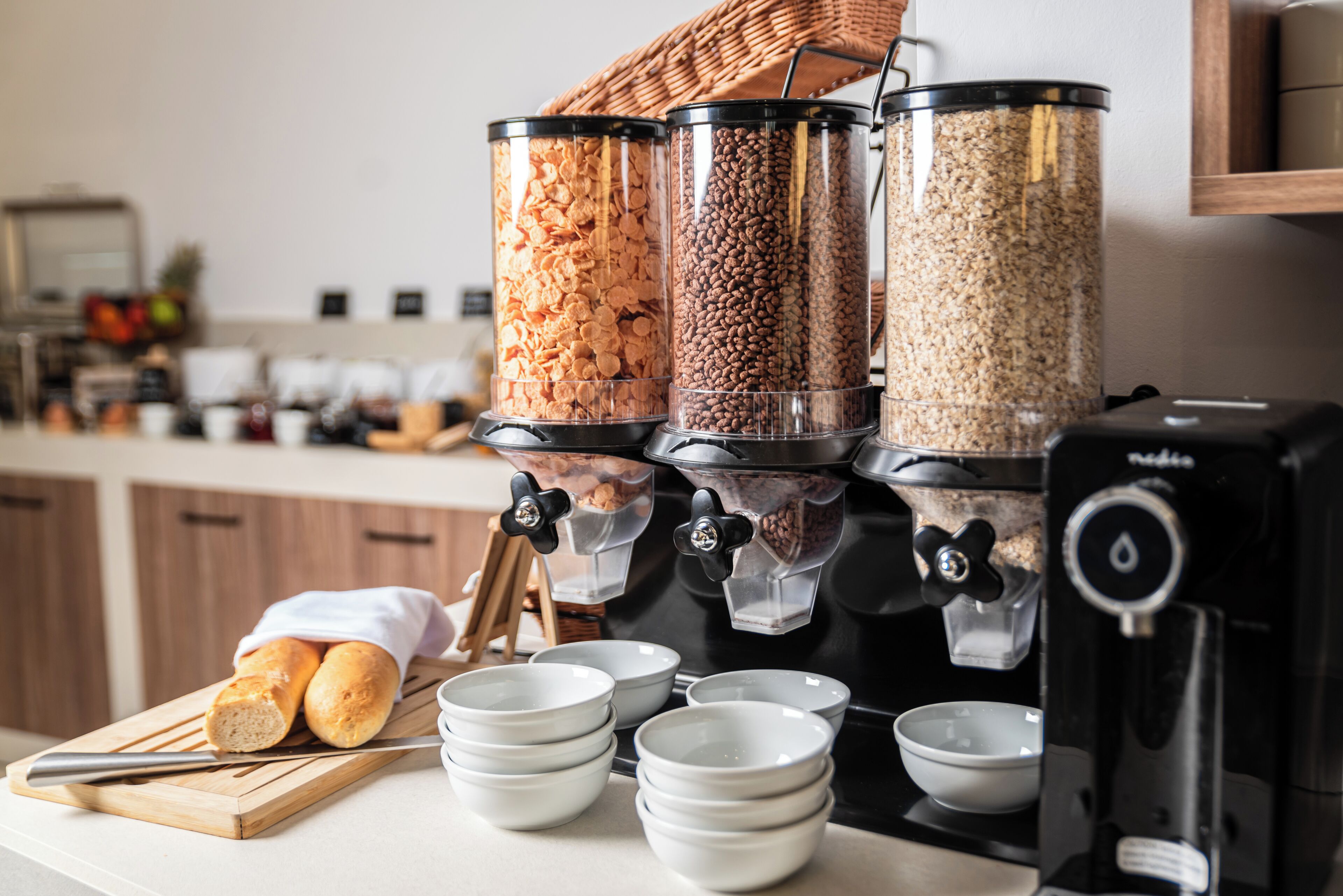 A coffee station with dispensers for coffee, tea, and creamer. There are bowls and a cutting board with bread.