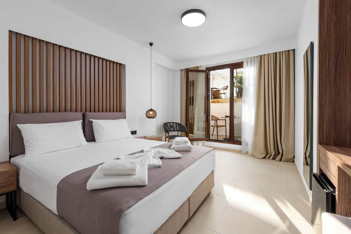 Habitación de hotel moderna con una cama grande, cabecera de madera, una mesita de noche, una silla y una puerta corrediza de vidrio que da a un espacio exterior.