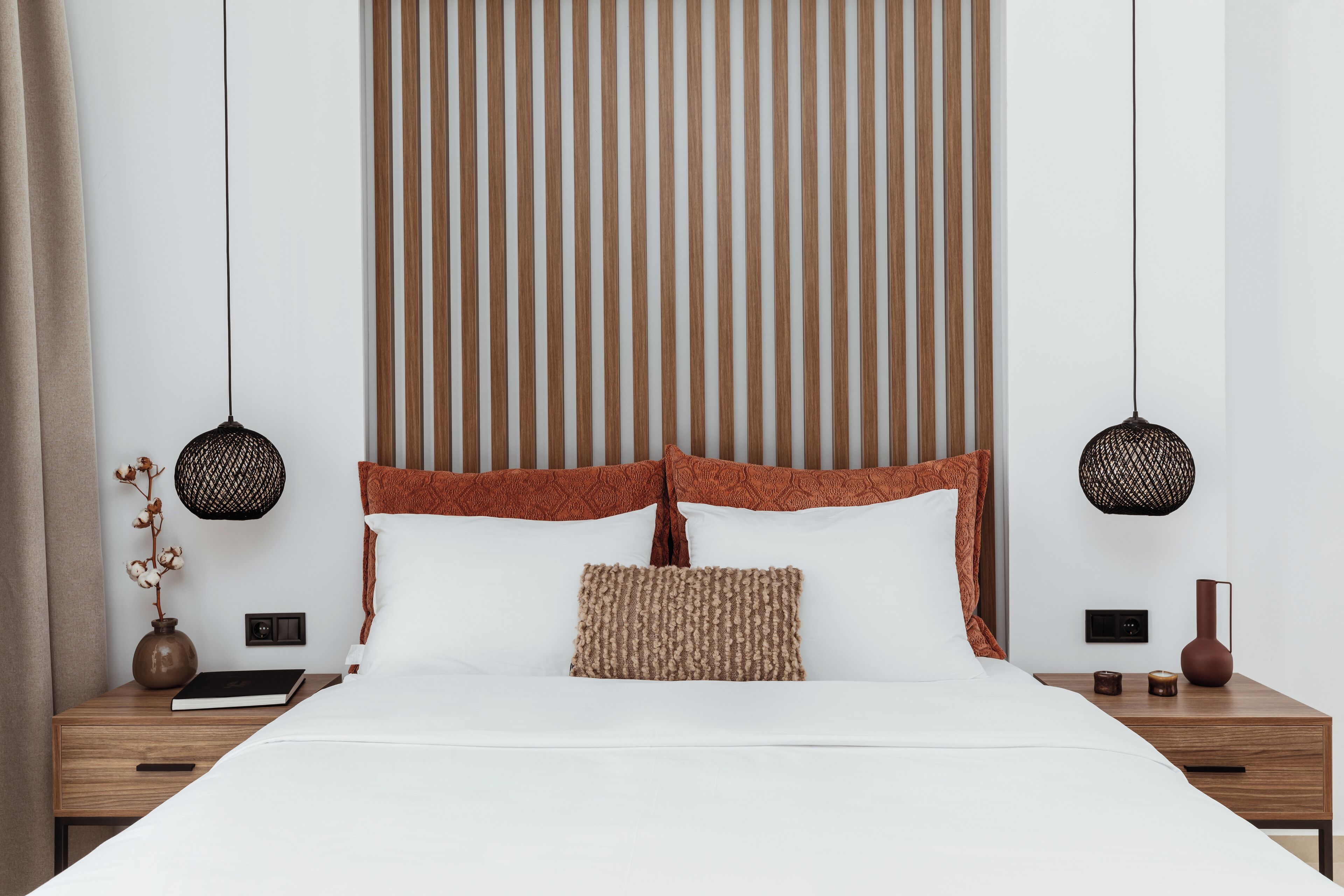 A neatly made bed with a wooden headboard, two bedside lamps, a vase with dried flowers, and a small table with a book and a remote control.