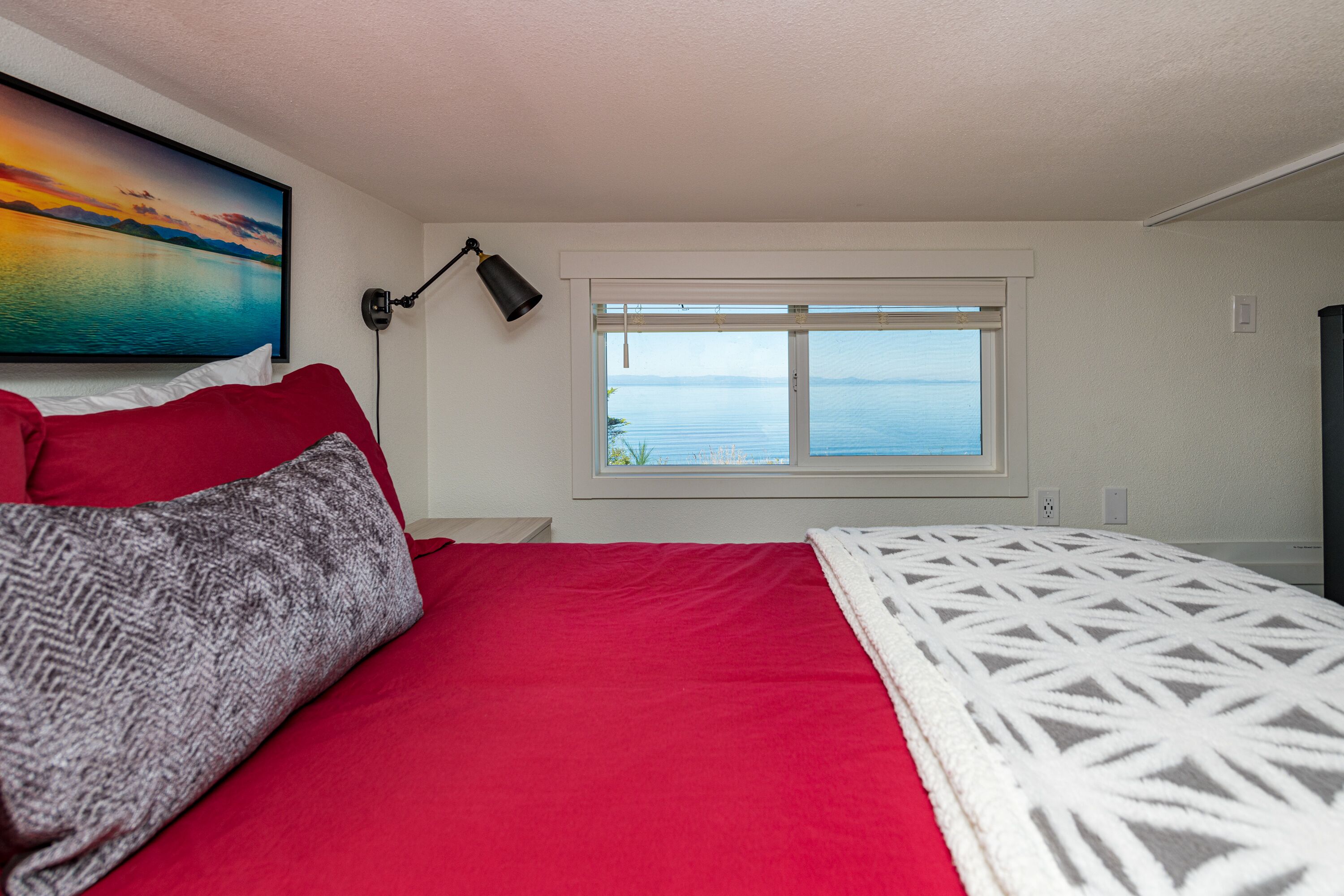 A bedroom with a bed, a window with a view of the sea, a painting on the wall, and a lamp.