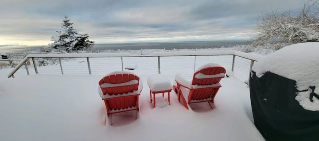 Um alpendre coberto de neve com duas cadeiras Adirondack vermelhas e uma pequena mesa.
