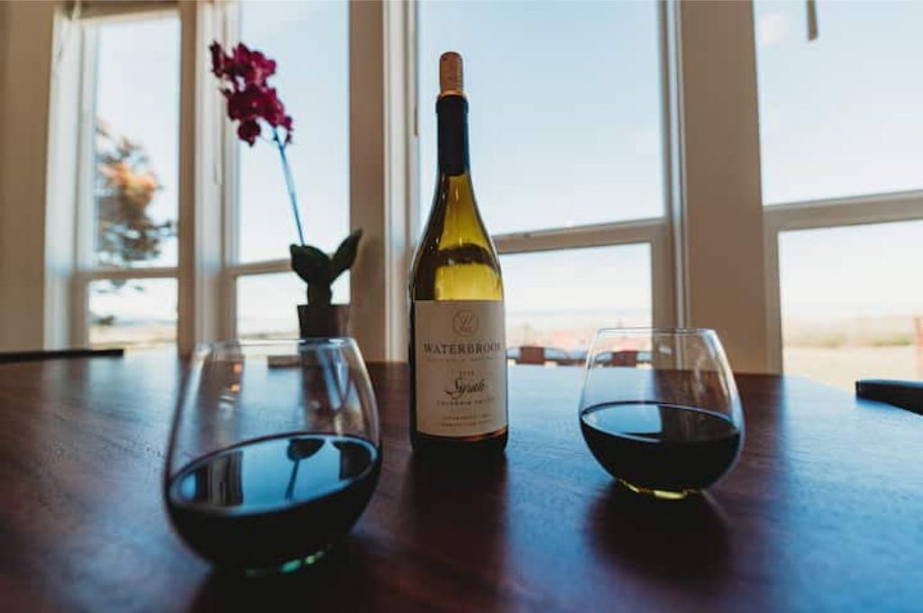 A bottle of Waterbrook Syrah wine, two wine glasses, and a vase with a flower on a wooden table by a window.