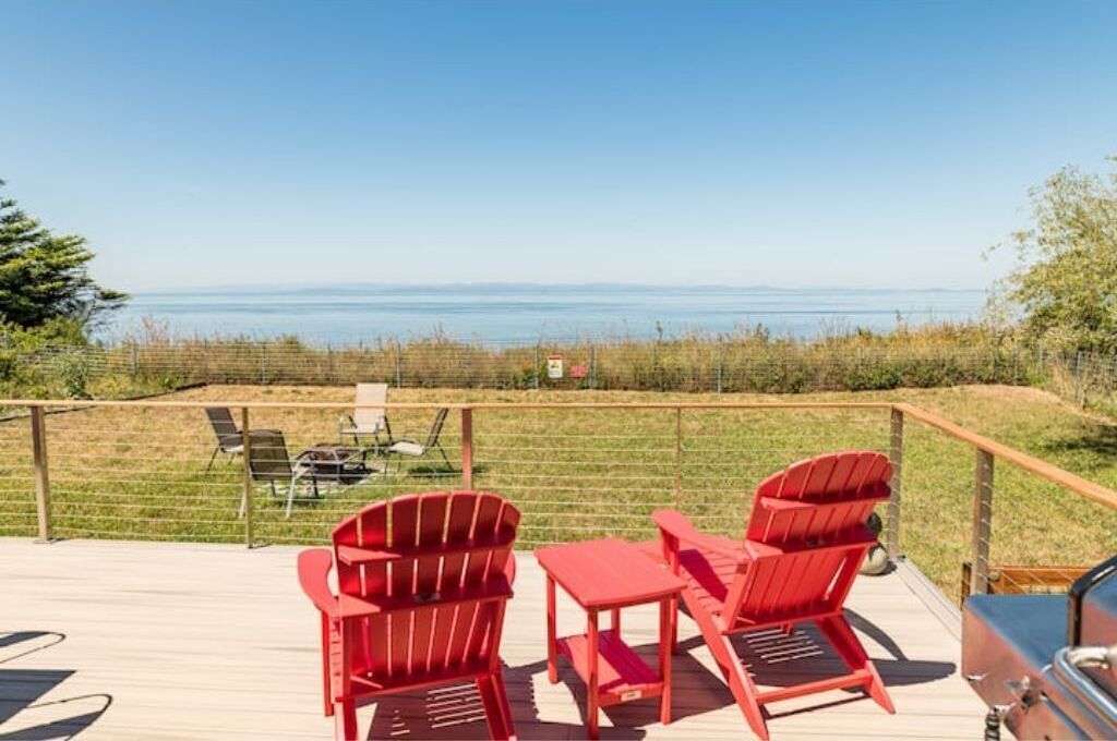Une terrasse avec des chaises Adirondack rouges et une table, donnant sur une zone herbeuse et un plan d’eau.