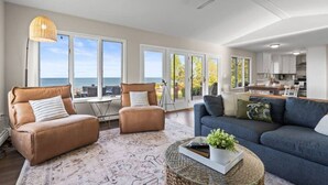 A spacious living area with a large window overlooking the sea, a ceiling fan, and a modern kitchen in the background. - Sheridan Shores Escape - Hot Tub w Amazing Views! (Lexington Township)