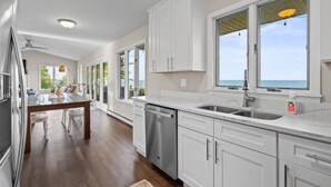 A modern kitchen with white cabinets, a stainless steel dishwasher, and a large window overlooking the ocean. - Sheridan Shores Escape - Hot Tub w Amazing Views! (Lexington Township)
