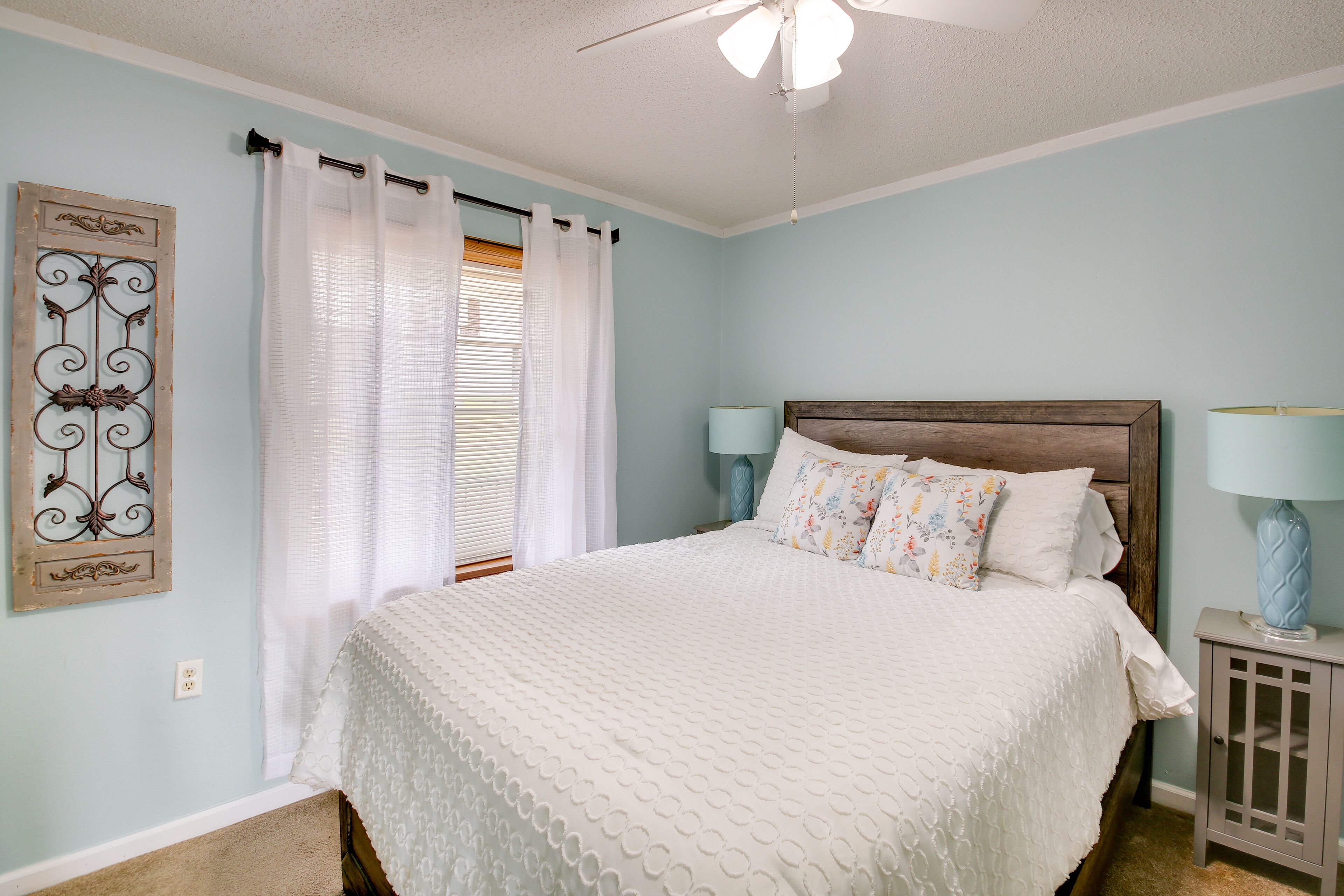 A bedroom with a bed, two bedside lamps, a window with curtains, and a wall-mounted decorative piece.