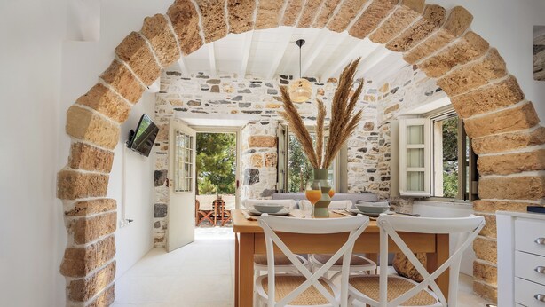 A cozy dining area with a stone archway, white chairs, and a wooden table. - Zos Suites (Naxos)
